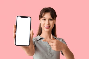 Gülümseyen genç bir kadın pembe arka plandaki boş cep telefonuna işaret ediyor.