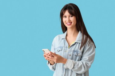 Gülümseyen genç kadın mavi arka planda cep telefonu kullanıyor.