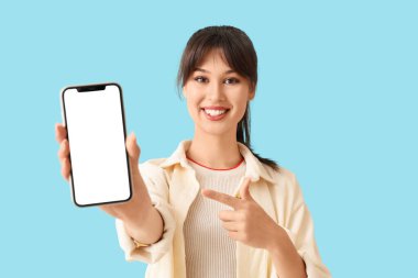 Gülümseyen genç bir kadın mavi arka plandaki boş cep telefonuna işaret ediyor.