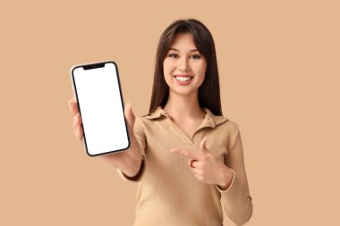 Gülümseyen genç bir kadın bej arka plandaki boş cep telefonuna işaret ediyor.