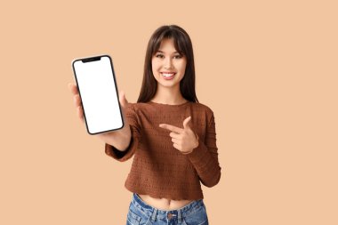 Gülümseyen genç bir kadın bej arka plandaki boş cep telefonuna işaret ediyor.
