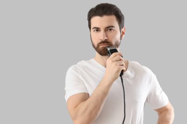 Yakışıklı genç adam sakalını gri arka planda elektrikli traş makinesiyle tıraş ediyor.