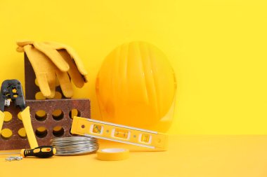 Sarı arka planda eldivenli, kalın şapkalı ve inşaat aletli tuğla