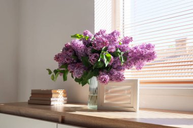 İçinde leylak çiçekleri, kitaplar ve odanın penceresinin yanındaki çekmecelerin çerçevesi olan vazo.