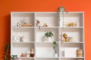 Çocuk odasında turuncu duvarın yanında oyuncakları ve bitkileri olan raf ünitesi.
