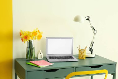 Modern dizüstü bilgisayar, güzel narsist çiçekler, lamba, kalemler ve defterler ışık duvarının yanındaki masada.