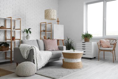 Konforlu kanepesi, göğüs çekmecesi, masası, çiçekleri, raf üniteleri, koltuğu ve sehpası olan bir oturma odası.