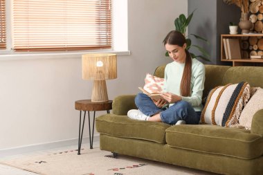 Oturma odasında kitapla kanepede oturan genç kadın.