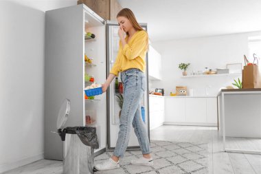 Mutfaktaki açık buzdolabının yanındaki çöp kutusuna bozuk mantar atan genç bir kadın.