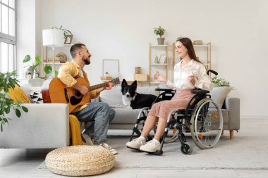Tekerlekli sandalyede, kocası ve köpeği evde gitar çalan genç bir kadın.
