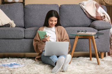 Dizüstü bilgisayarla çalışan güzel genç bir kadın ve oturma odasındaki halıda not defteri.
