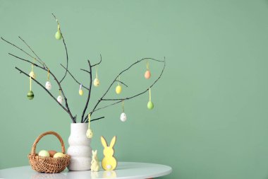 Ağaç dallı vazo, Paskalya yumurtası, tavşanlar ve sepet yeşil duvarın yanındaki masada, yakın plan.
