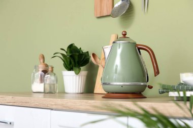 Mutfaktaki yeşil duvarın yanındaki masada fincanlı elektrikli çaydanlık ve çatal bıçak.