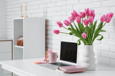 Modern dizüstü bilgisayarlı şık çalışma alanı, bir fincan çay ve beyaz tuğla duvarın yanında güzel lale çiçekleri.