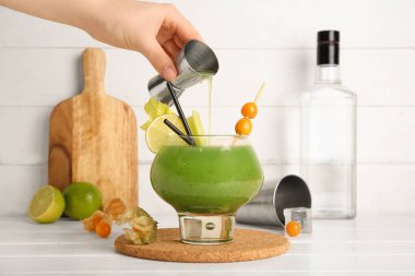 Beyaz ahşap arka planda yeşil kanlı Mary kokteyli bardağına votka döken bir kadın.