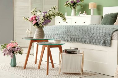 Yatak odasının içinde masa, dergi tutacağı ve güzel çiçekler var.