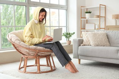 Evdeki koza sandalyesinde dizüstü bilgisayar kullanan genç bir kadın.
