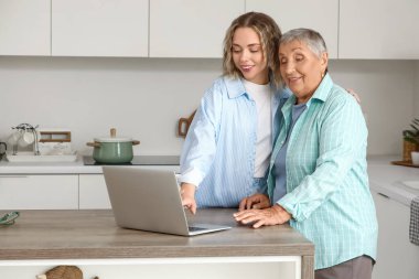 Torunuyla birlikte mutfaktaki masada dizüstü bilgisayar kullanan yaşlı bir kadın.