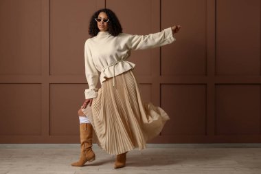 Kahverengi duvarın yanında güneş gözlüklü Afro-Amerikan bir kadın.