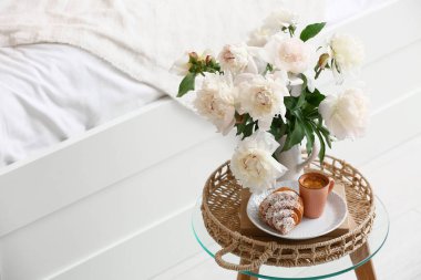 Şakayık çiçekli vazo, bir fincan kahve, kruvasan ve yatak odasındaki masada kitaplar.