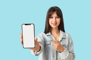 Gülümseyen genç bir kadın mavi arka plandaki boş cep telefonuna işaret ediyor.