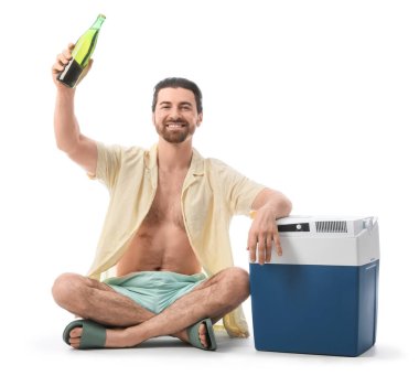 Portatif buzdolabı ve beyaz arka planda izole edilmiş soğuk bira şişesiyle oturan mutlu genç adam, yakın plan.