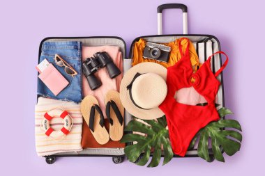 İçinde giysiler, mayolar, plaj aksesuarları ve eflatun arka planda dürbünler olan valizi aç.