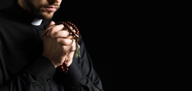 Genç rahip karanlık arka planda boncuklarla dua ediyor. Mesaj için yer var, yakın plan.