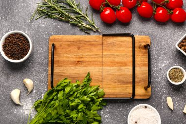 Ahşap mutfak tahtası, taze otlar, domatesler ve arkaplanda baharatlar.