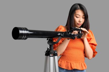 Genç Asyalı kadın gri arka planda teleskop kullanıyor.