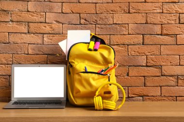 Kahverengi duvarın yanındaki ahşap masada kırtasiye, dizüstü bilgisayar ve kulaklık bulunan sarı okul çantası.