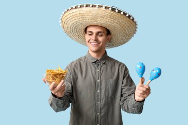 Marakas ve mavi arka planda bir kase nachos ile sombrero giyen yakışıklı genç adam.