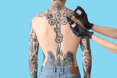 Dövmeci, dövme spreyi ve krem tutuyor. Mavi arka planda dövmeli bir adamın yanında.