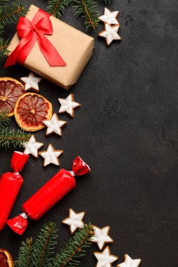 Lezzetli yıldızlı kompozisyon şekilli Noel kurabiyeleri, şekerler, turuncu kurumuş parçalar, hediye kutusu ve koyu arkaplanda köknar ağacı dalları