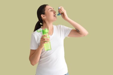 Yeşil arka planda astım spreyi kullanan, su şişeli genç bir kadın.