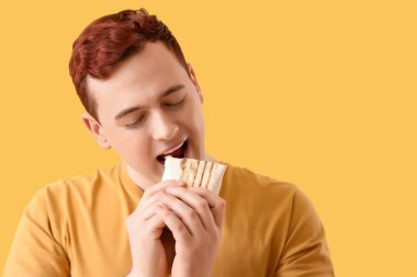 Young man eating tasty doner kebab on yellow background