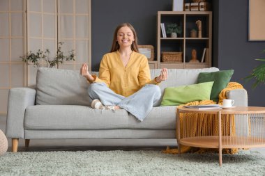 Genç bir kadın oturma odasındaki gri kanepede meditasyon yapıyor.