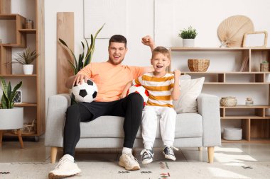 Genç adam ve küçük oğlu evde futbol toplarıyla futbol takımı için tezahürat yapıyorlar.