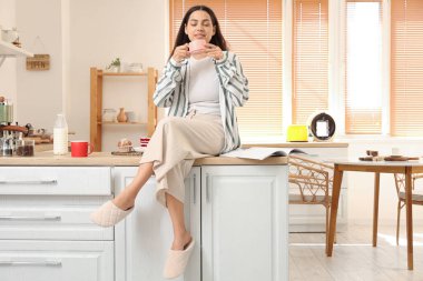 Genç, mutlu bir kadın, elinde bir fincan taze kahve ve mutfakta tatlı okuyarak.