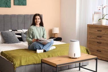 Yatak odasındaki modern nemlendiricinin yanında rahat bir yatakta dergi okuyan genç bir kadın.