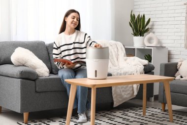 Genç kadın kanepede oturuyor ve oturma odasındaki modern nemlendiriciyi açıyor.