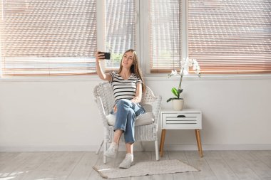 Orkide çiçeğinin yanındaki koltukta otururken selfie çeken genç bir kadın.