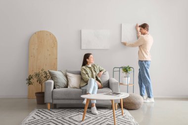 Genç adam ışık duvarına resim asıyor ve karısı evde.