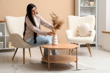 Güzel genç bir kadın, şık oturma odasındaki beyaz koltuğun yanındaki vazoya çim koyuyor.