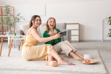 Pedikür için oje örnekleri taşıyan genç kadınlar oturma odasında selfie çekiyorlar.