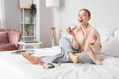 Ayak parmağı ayırıcı ve ojeli mutlu genç bir kadın yatak odasında pedikür yapıyor.