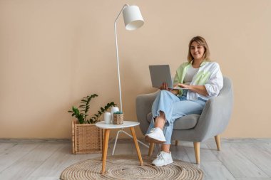 Evdeki bej duvarın yanındaki koltukta dizüstü bilgisayar kullanan genç bir kadın.