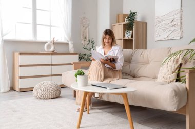 Evdeki kanepede dergi okuyan genç bir kadın.