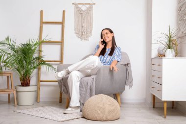 Evdeki koltukta otururken cep telefonu kullanan genç bir kadın.