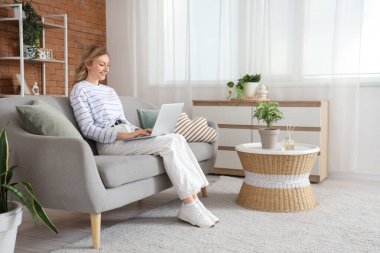 Evdeki kanepede dizüstü bilgisayar kullanan güzel genç bir kadın.